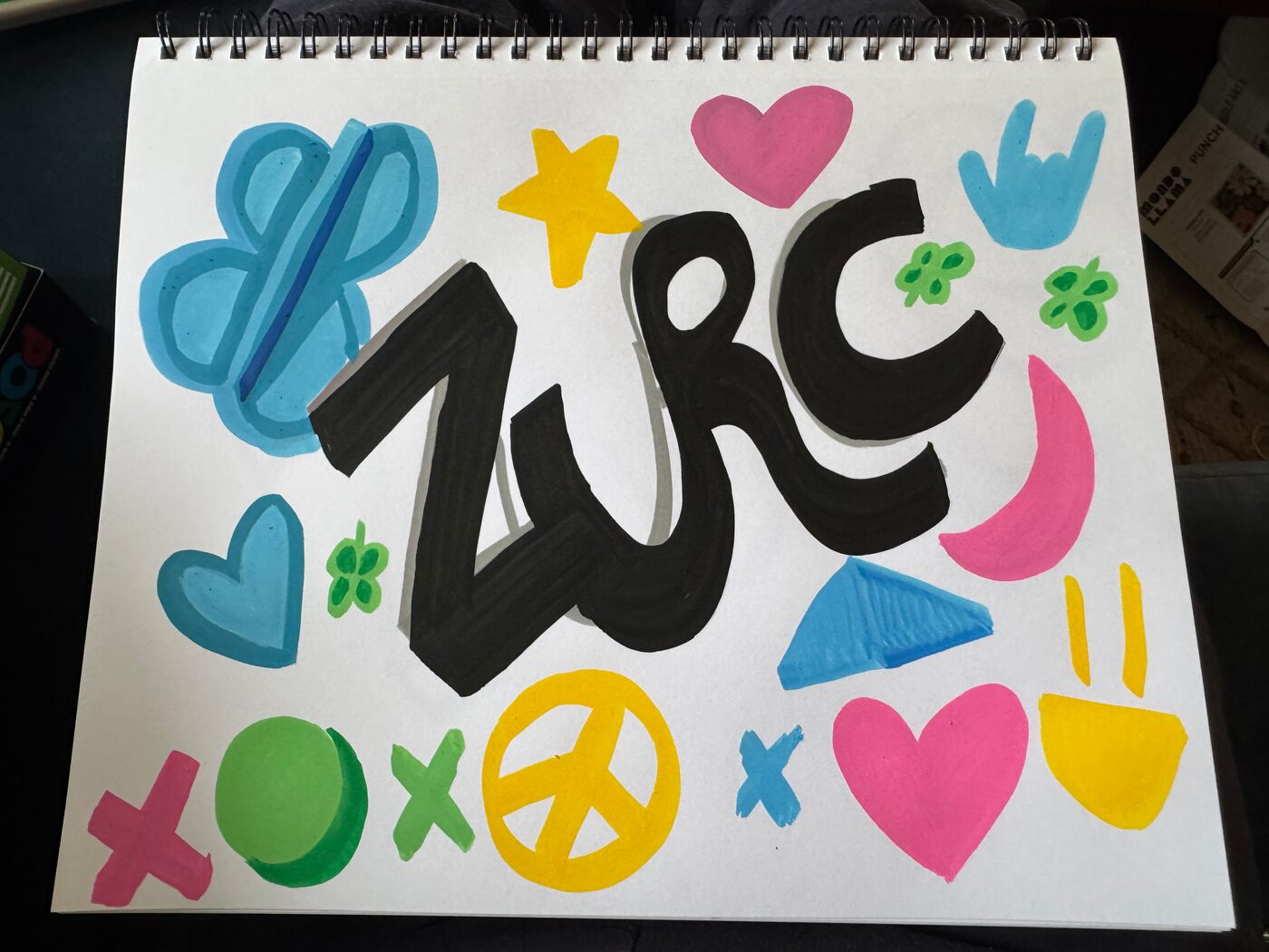 ZURC in thick black marker with gray shadows across a sketchbook page, ringed by pink and blue hearts, a yellow star, a yellow peace sign and a yellow smiling face, green clovers, x's, a pink crescent and a blue hand sign